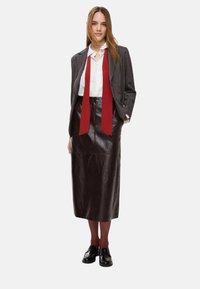 Blazer a quadri grigio sopra una camicia bianca, abbinato a una gonna midi in pelle marrone e scarpe nere. La cravatta rossa aggiunge un accento a contrasto.