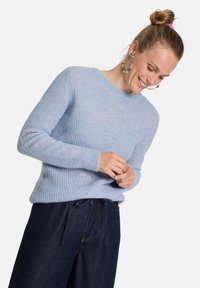 Hellblauer Strickpullover mit Rundhalsausschnitt, gerippter Textur und dekorativen Stichdetails. Kombiniert mit dunklen Jeans.