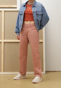 Veste en denim bleu clair sur une chemise cropped couleur rouille, associée à un jean rose coupe droite et des baskets blanches, le tout sur un fond neutre.