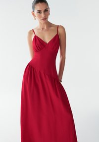 Vestido maxi rojo con tirantes finos y un corsé ajustado. Presenta costuras diagonales y tejido fruncido en el dobladillo. Textura suave.