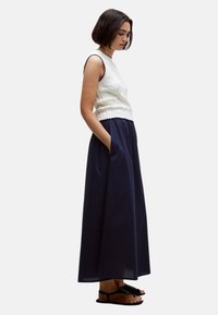 Top sin mangas de punto blanco con bordes ondulados, combinado con una falda larga azul marino con bolsillos laterales. Sandalias planas negras completan el look.