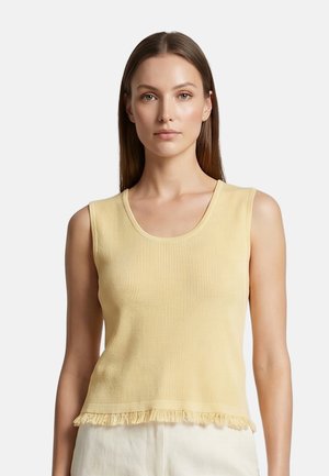 Donna che indossa un top a coste giallo chiaro senza maniche con dettagli di frange all'orlo e pantaloni chiari su uno sfondo uniforme.