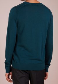 Teal knit sweater with long sleeves, crew neckline, ribbed hem, and side slit details, worn over black pants. Soft texture and fitted design.