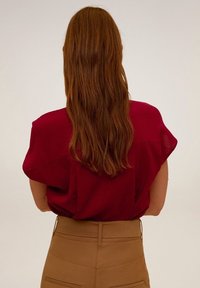 Blouse bordeaux à manches courtes avec une coupe ample, vue de dos. Des cheveux longs et ondulés couvrent partiellement l'épaule. Pantalon taille haute couleur sable.