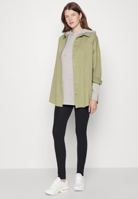 Túlméretezett olíva zöld gombos ing egy szürke bordás kapucnis pulóver fölött, fekete leggings-szel és fehér sneakers-szel párosítva.