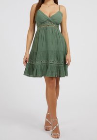 Un vestido de verano de tela verde con un corpiño ajustado, dobladillo con volantes, paneles texturizados y acentos de encaje. Tirantes de espagueti ajustables y material ligero.