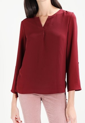 Kvinde iført en langærmet vinrød bluse med delt halsudskæring og lyserøde fløjlsbukser mod en hvid baggrund.