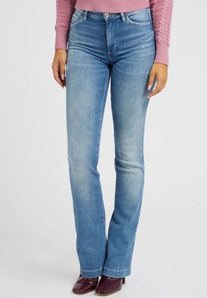 Kvinde iført lyseblå jeans med lige ben, lyserød striktrøje og mørkelilla hælede sko, der står mod en hvid baggrund.
