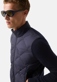 Veste sans manches matelassée navy avec texture en losanges et col montant ; portée sur un haut à manches longues en tricot bleu foncé ; complétée par des lunettes de soleil noires.