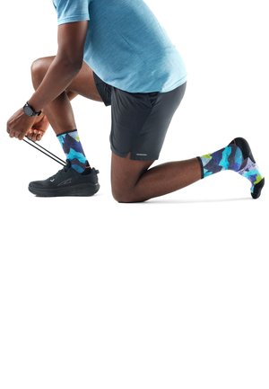 Hombre arrodillado en una rodilla atándose los cordones de las zapatillas negras para correr, vistiendo una camiseta deportiva azul, pantalones cortos negros, calcetines con patrones coloridos y un reloj inteligente.