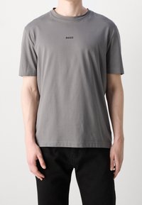 Grå bomullströja med korta ärmar, rund halsringning och en liten svart "BOSS"-logotyp på bröstet, kombinerad med mörka jeans.