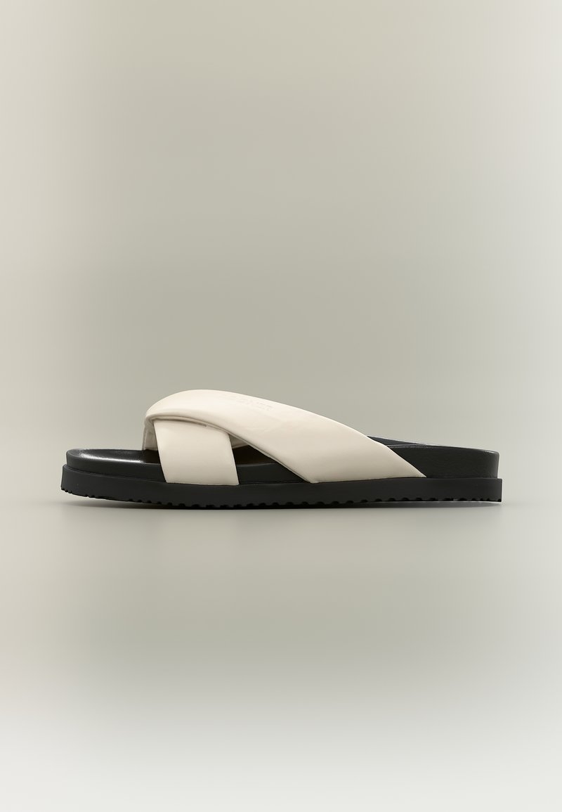 Sandale mule à semelle noire avec larges brides croisées en cuir blanc cassé et semelle extérieure texturée, vue de profil sur un fond uni.