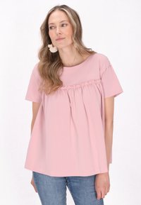 T-shirt rosa chiaro a maniche corte con cucitura arricciata sotto il busto, design fluido, texture morbida, abbinato a jeans azzurri.