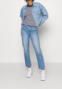 Veste en denim bleu clair sur une chemise rayée bleu marine et blanche, associée à un jean bleu clair et des baskets blanches. Design simple et décontracté.