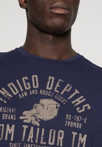 Camiseta de algodón azul marino con un estampado gráfico que incluye un diseño de cámara y texto en color beige claro. Cuello redondo y mangas cortas.