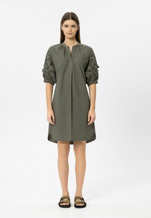 Woman standing wearing olive green knee-length dress with floral cutout puff sleeves and brown sandals with buckles.