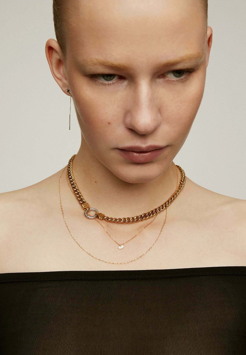 Collar de cadena de oro con un cierre redondo, superpuesto con delicadas cadenas de oro y un pequeño encanto, llevado sobre una blusa negra sin hombros.