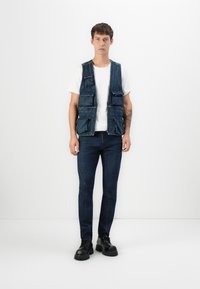 Denim prsluk s više prednjih džepova, nosi se preko bijele majice. Model ga kombinira s tamnoplavim trapericama i crnim čizmama s debelim potplatom.