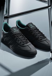 Baskets basses noires avec une doublure intérieure turquoise, logo AMG sur la languette, placées sur une surface réfléchissante près d'un cadre chromé.