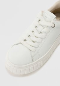 Baskets en cuir blanc avec semelle épaisse texturée et lacets blancs duveteux, inclinées pour montrer les détails avant et de côté.