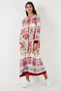 Naine, kes kannab pikka valget lillelist kleiti punaste, roosade ja roheliste mustritega, valgete tossudega, taustaks tavaline sein.