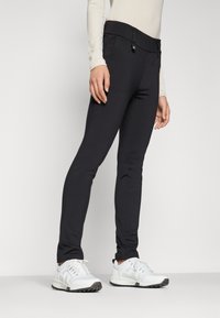 Pantaloni neri aderenti con una cintura larga e dettagli cuciti. Il materiale sembra elastico. Abbinati a scarpe da ginnastica bianche.