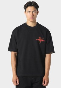 Jonge man met donker haar, gekleed in een eenvoudig zwart oversized T-shirt met een kleine rode afbeelding en tekst op de linkerkant van de borst, staand tegen een lichte achtergrond.