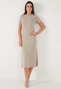 Beige, short-sleeved, midi dress made of smooth fabric, featuring side slits for movement. Model poses with hand on hip, wearing sandals.
