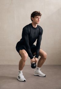 Črna športna majica z dolgimi rokavi, črne kratke hlače in bele teniske; drži črno kettlebell, na nevtralnem ozadju.