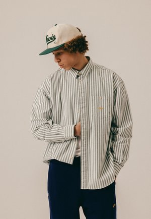 Jeune personne aux cheveux bouclés portant une casquette blanche et verte, une chemise à boutons grise et blanche à rayures, et un pantalon sombre, regardant vers le bas et sur le côté.