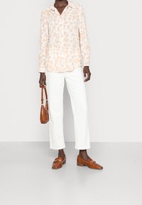 Blouse pêche clair avec un motif abstrait blanc, pantalon blanc, mocassins marron et un petit sac à main marron texturé.