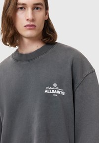 Szary sweatshirt z okrągłym dekoltem i ściągaczami, z białym tekstem i logo. Miękka tkanina o lekko zużytej fakturze.