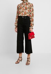 Schwarze hochgeschnittene weitbeinige Jeans, kombiniert mit einer langärmeligen Bluse im floralen Muster und einer kleinen roten Handtasche. Riemchensandalen mit Absatz in Schwarz.