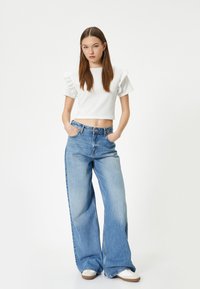 Hvid ribbet cropped top med flæseærmer, parret med lyseblå cropped jeans. Modellen står med hænderne i lommerne og har hvide sneakers på.