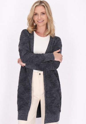 Femme souriante aux cheveux blonds portant un cardigan gris foncé ouvert sur un haut blanc et un pantalon beige clair, debout les bras croisés.