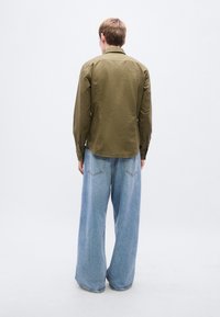 Πουκάμισο λαδί με γιακά και μανσέτες με κουμπιά, σε συνδυασμό με ανοιχτό μπλε τζιν παντελόνι φαρδιάς γραμμής, με πίσω τσέπες και χαλαρή εφαρμογή.