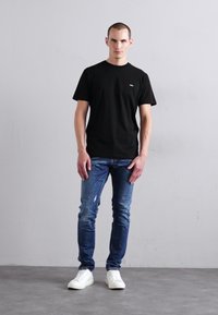 Black cotton t-shirt with short sleeves, paired with distressed blue jeans. White sneakers complete the outfit, against a neutral background.
