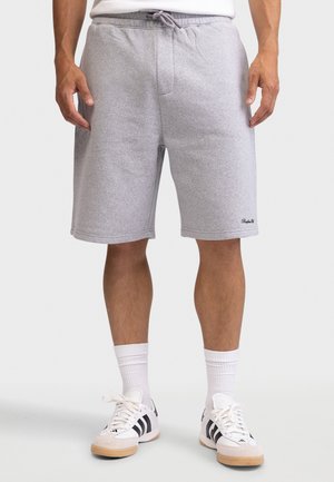 Άνδρας που φοράει ανοιχτό γκρι σορτς με κορδόνι, λευκές κάλτσες μέχρι τον αστράγαλο και λευκά αθλητικά παπούτσια με μαύρες ρίγες, στέκεται σε απλό φόντο.