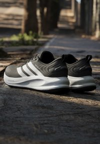 Paire de chaussures de course Adidas noires et blanches posée sur un pavement extérieur irrégulier avec une lumière douce et un arrière-plan ombragé.