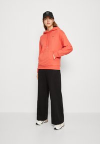 Felpa corallo con tasca a marsupio e cordino, abbinata a pantaloni larghi neri e sneakers bianche con dettagli grigi. Cappello nero in testa.