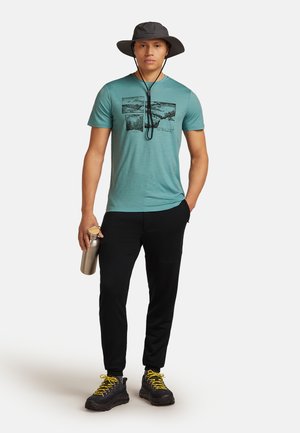 Mand iført en teal grafisk t-shirt, sorte bukser, en grå bredskygget hat og vandrestøvler med gule snørebånd, mens han holder en metal vandflaske.