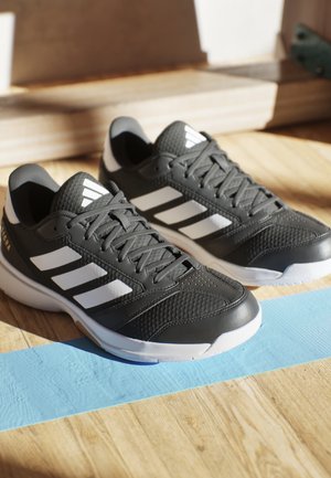 Schwarz-weiße Adidas Sportschuhe mit Schnürsenkeln auf einem Holzboden mit blauer Klebebandlinie im Sonnenlicht.