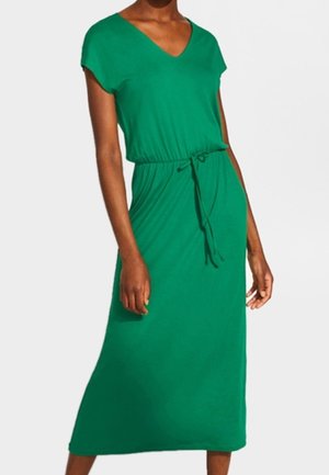 Femme portant une robe midi verte sans manches avec un décolleté en V et une taille à cordon, debout devant un fond blanc uni.