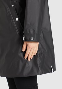 Černý nepromokavý plášť proti dešti s lesklým povrchem, vybavený přední kapsou, bílými patentkami a staženým zápěstím s kulatým logem.