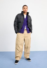 Giacca a vento nera con colletto alto, cardigan viola e pantaloni cargo beige. Sneakers nere completano l'outfit, che presenta una vestibilità casual.