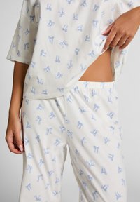 Lichtgewicht katoenen pyjama set met een losvallend topje en broeken met elastische taille, beiden versierd met kleine blauwe strikpatronen op een witte achtergrond.