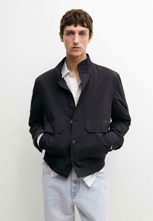 Jeune homme aux cheveux bruns courts portant une veste noire boutonnée sur une chemise blanche et un jean bleu clair, debout les mains dans les poches.