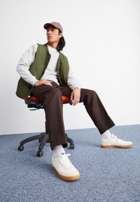 Mężczyzna w różowej czapce, zielonej kamizelce, szarym swetrze, brązowych spodniach i białych trampkach Nike siedzi na obrotowym krześle biurowym na niebieskim dywanie.