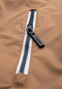 Braunfarbener Jackenstoff mit einem diagonalen Reißverschluss, der einen schwarzen Ziehgriff und marineblaue Zähne aufweist, umrahmt von einem weißen Band entlang der Öffnung.