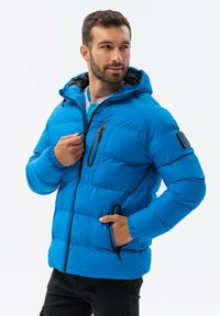 Modrá zateplená bunda s kapucí, vybavená černými zipy a akcenty. Obsahuje boční kapsy a stylový prošívaný design.